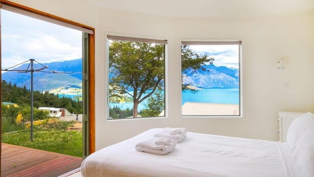 A bedroom with a bed, two windows with mountain and lake views, and a balcony with a wooden deck. - Ruby's Fernhill Vista (Queenstown)