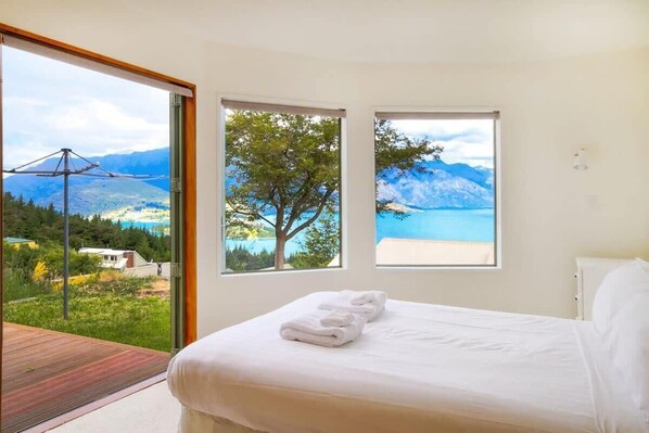 A bedroom with a bed, two windows with mountain and lake views, and a balcony with a wooden deck.