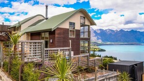 A modern house with a green roof, wooden siding, and large windows. It is situated near a lake with mountains in the background. - Ruby's Fernhill Vista (Queenstown)