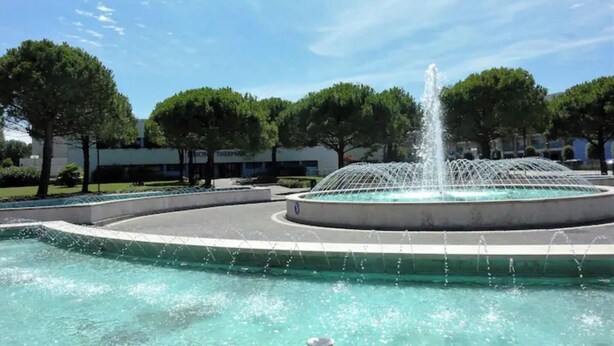 A circular fountain with water jets in the center, surrounded by a paved area with trees and buildings in the background.