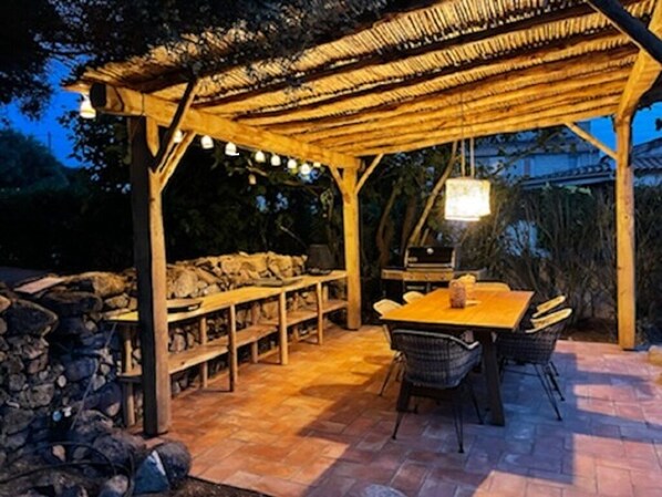 Un patio exterior con una pérgola de madera, asientos de piedra y una mesa con sillas.