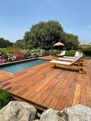 Una terraza de madera con piscina, sillas de descanso y un quiosco de techo de paja.
