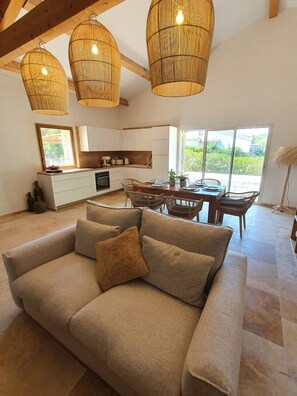 Una sala de estar moderna con un sofá color beige, una zona de comedor con sillas de mimbre y una cocina con armarios blancos y un ventanal grande.
