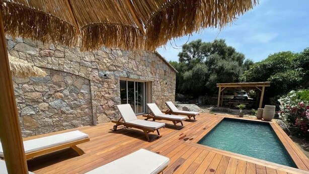 A wooden deck with lounge chairs, a swimming pool, and a stone building with a thatched roof.