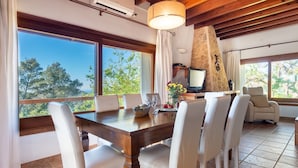 Un comedor con mesa de madera y sillas blancas, chimenea de piedra y ventanas amplias con vista a árboles.