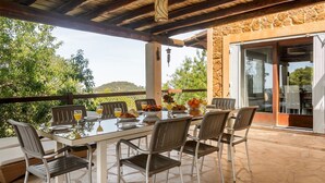 Un área de comedor al aire libre con una pared de piedra, techo de madera y vistas a zonas verdes y colinas.
