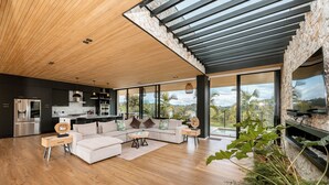 A modern living room with a wooden ceiling, large windows, a sofa, and a kitchen in the background.