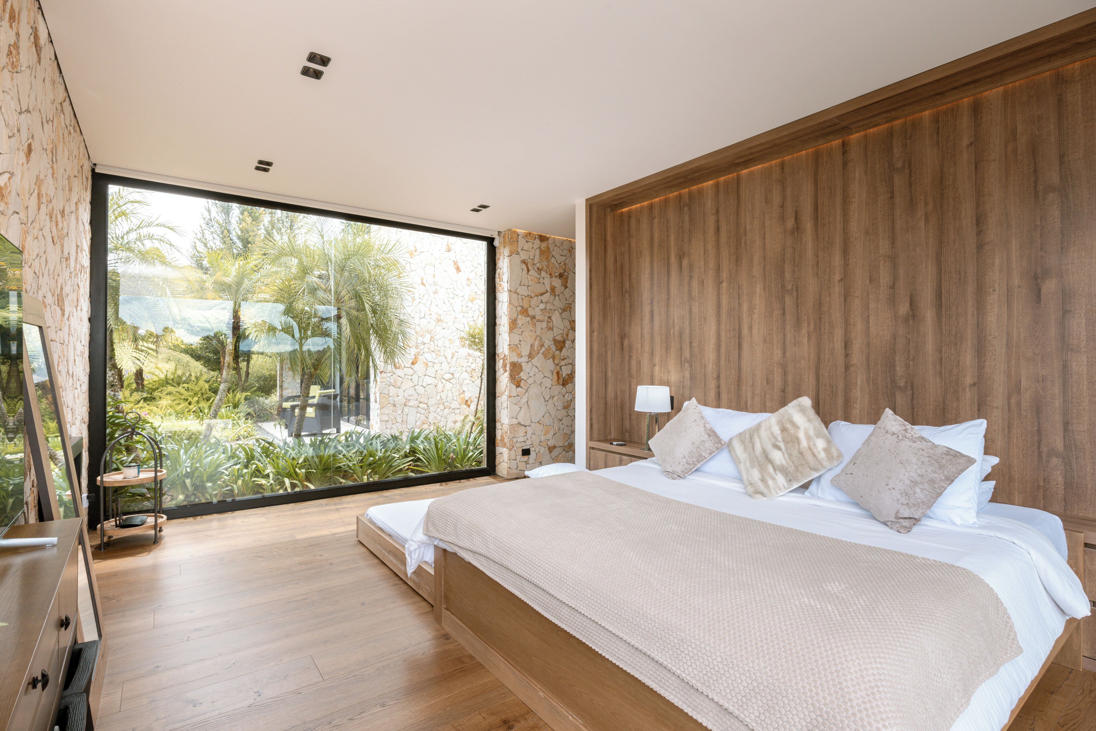 A modern bedroom with a large bed, wooden headboard, and a view of greenery through a glass door.