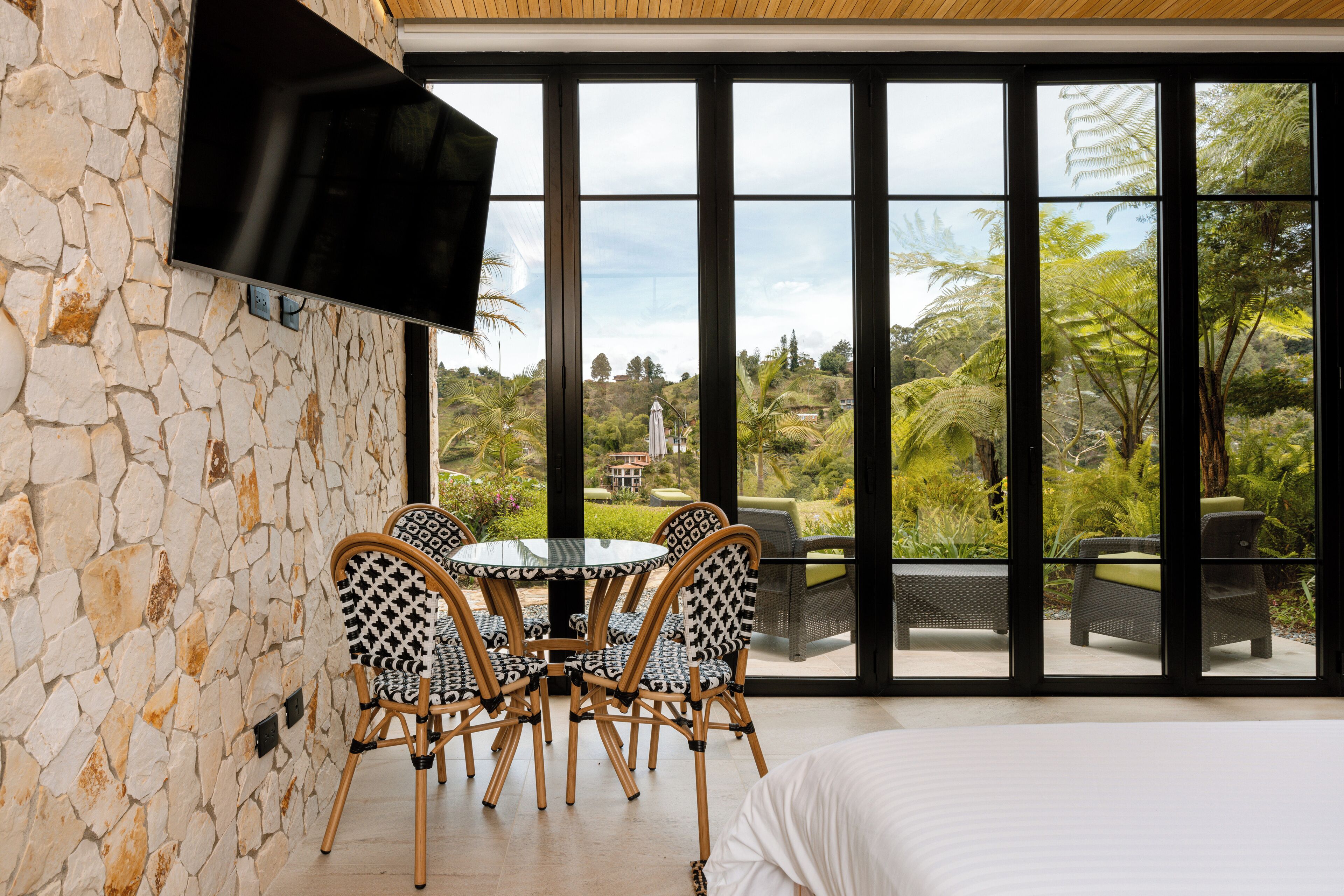 A room with a stone wall, a flat-screen TV, a round table with chairs, and a bed.