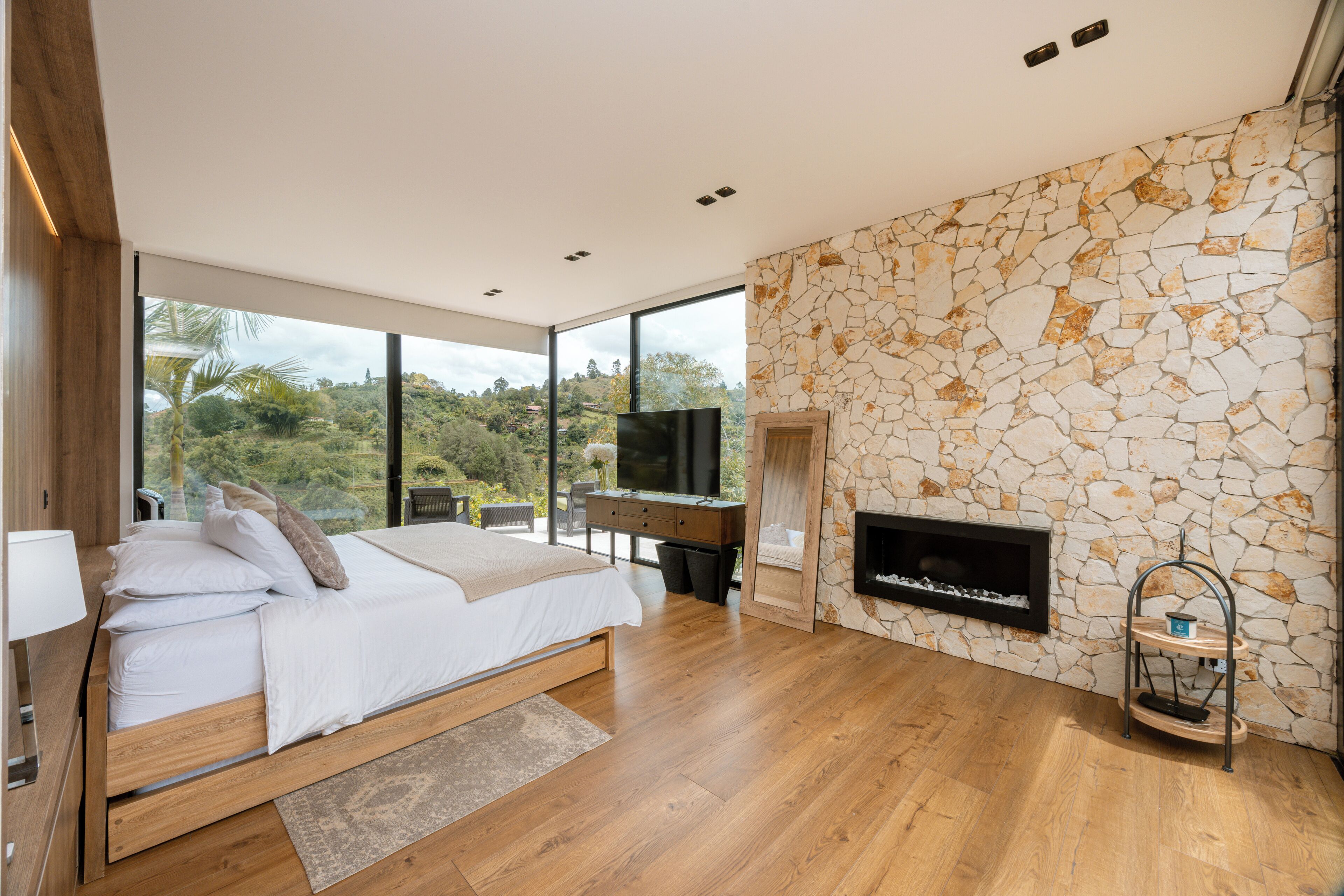 A modern bedroom with a large bed, a stone wall, a fireplace, and a TV.