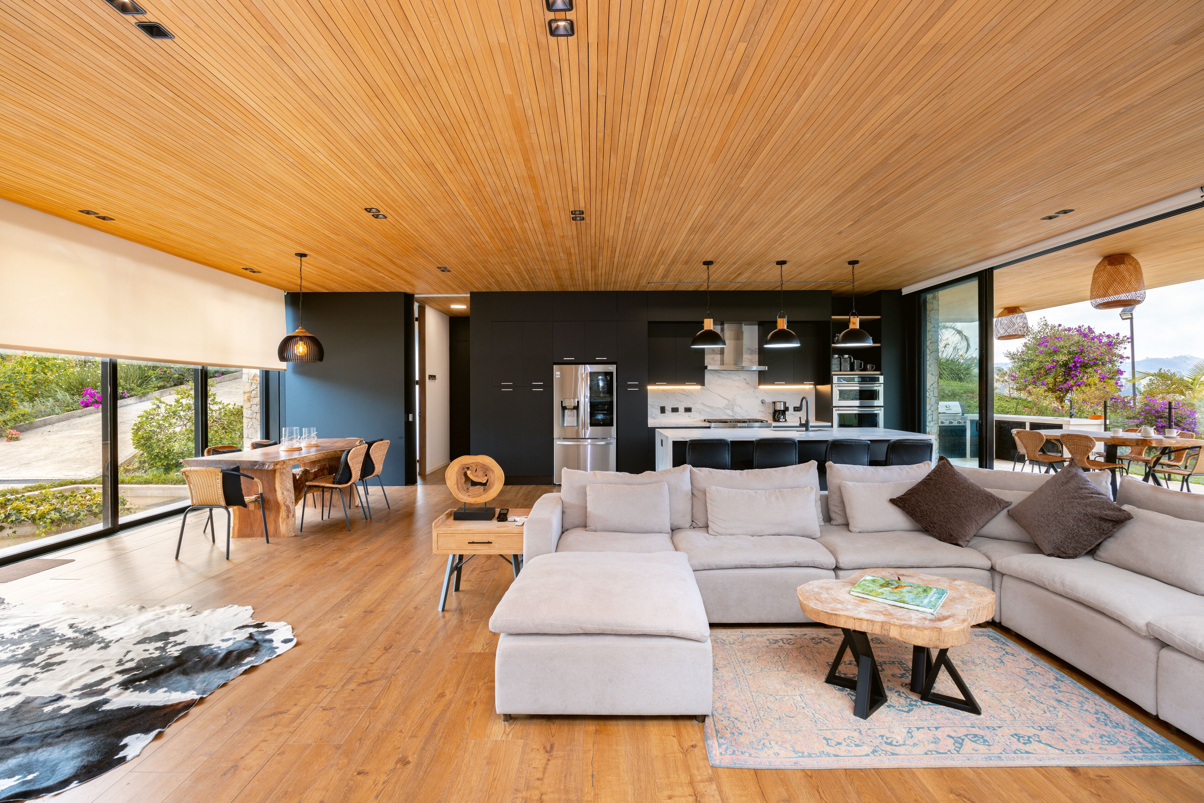A modern living room with a wooden ceiling, a large sectional sofa, a dining area, and a kitchen with a large island.