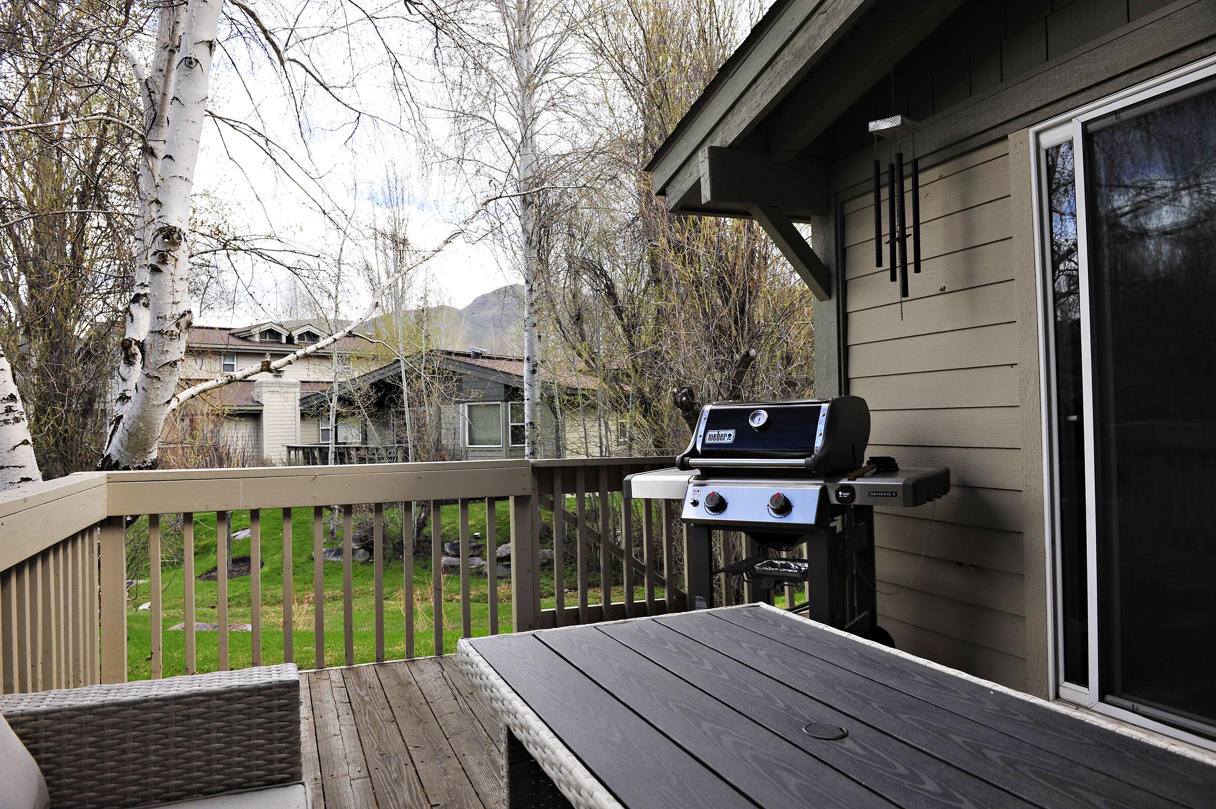 A deck with a grill, a table, and a view of a backyard with trees and houses.