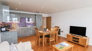 A modern living room with a sofa, a wooden dining table with chairs, a television, and a kitchen area with cabinets and a countertop. - Sandsend View (Whitby)