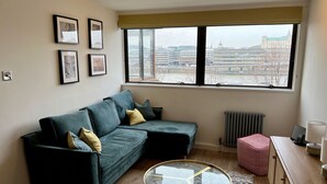 Ein modernes Wohnzimmer mit einem grünen Sofa, einem Glastisch und Blick auf eine Stadtlandschaft durch große Fenster.