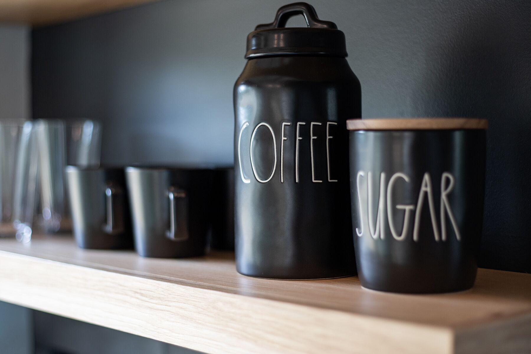 Una fila di contenitori neri con le scritte 'COFFEE' e 'SUGAR', posti su un ripiano di legno.