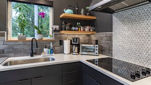 A modern kitchen with a built-in sink, a coffee machine, a toaster, and a window with plants. - Kitty Hawk Reserve OBX Tiny House! Hot Tub & Beach included! (Kitty Hawk)