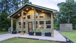 A two-story wooden cabin with multiple windows, a covered porch, and outdoor seating. - Kitty Hawk Reserve OBX Tiny House! Hot Tub & Beach included! (Kitty Hawk)