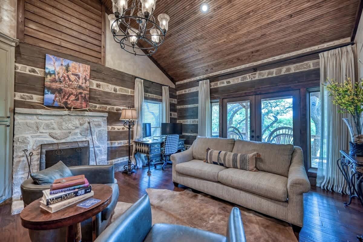 A log cabin interior with a stone fireplace, a sofa, a round table with books, and a chandelier.