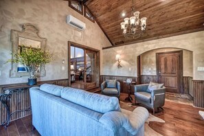 A spacious living room with a high ceiling, a large sofa, two armchairs, a chandelier, and a potted plant.