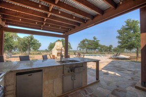 An outdoor kitchen area with a stone countertop, built-in sink, and storage cabinets, surrounded by stone flooring and outdoor furniture. - Lux Ranch w/Pool-Hot-Tub Near Fred! (Fredericksburg)