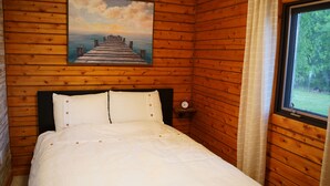 A wooden-paneled room with a bed, a painting of a pier, and a window with curtains. - Cozy lakeside cottage with breathtaking views & amazing fishing  (Yarker)