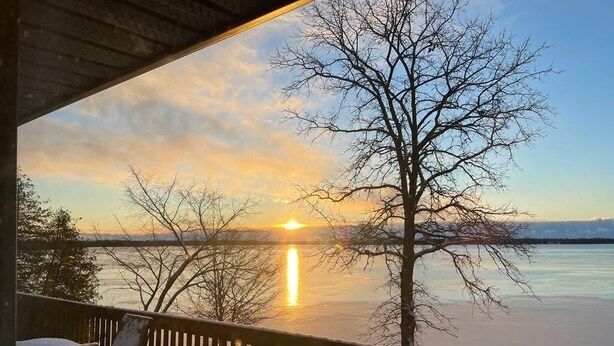 A deck with a view of a sunset over a body of water, a bare tree, and a chair. - Cozy lakeside cottage with breathtaking views & amazing fishing  (Yarker)