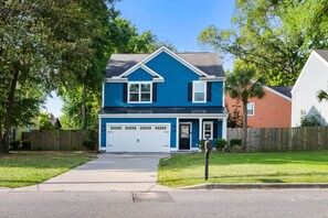 A two-story house with a blue exterior, a white garage door, and a front yard with a sidewalk leading to the entrance. - Private Backyard | Park Circle | The Sandy Palm! (North Charleston)