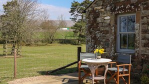 A stone building with a small patio table and two chairs, a window, and a view of a fenced field with trees. - Herdie Hideaway (Appleby-in-Westmorland)