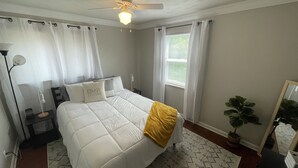 Ein Schlafzimmer mit einem Bett, Deckenventilator, Nachttisch, Fenster mit Vorhängen und einer Zimmerpflanze.