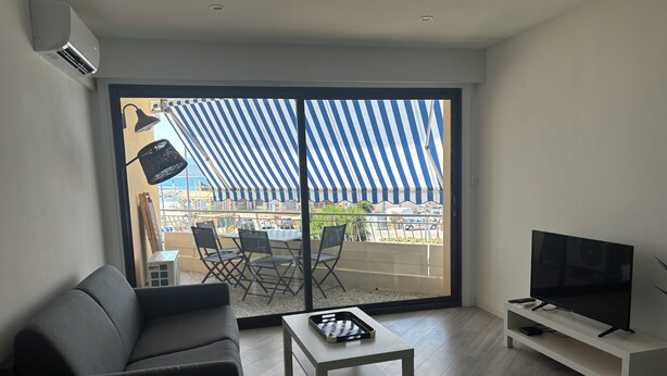 A modern living room with a grey sofa, a white coffee table, and a TV on a white stand. A large sliding glass door leads to an outdoor area with a striped awning and a table with chairs. - Beautiful new 2-room apartment, Saint-Raphael golf course, close to beaches (Saint-raphael)