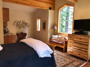 Quarto com cama, penteadeira de madeira, televisão, janela com vista para árvores e luminária na parede.