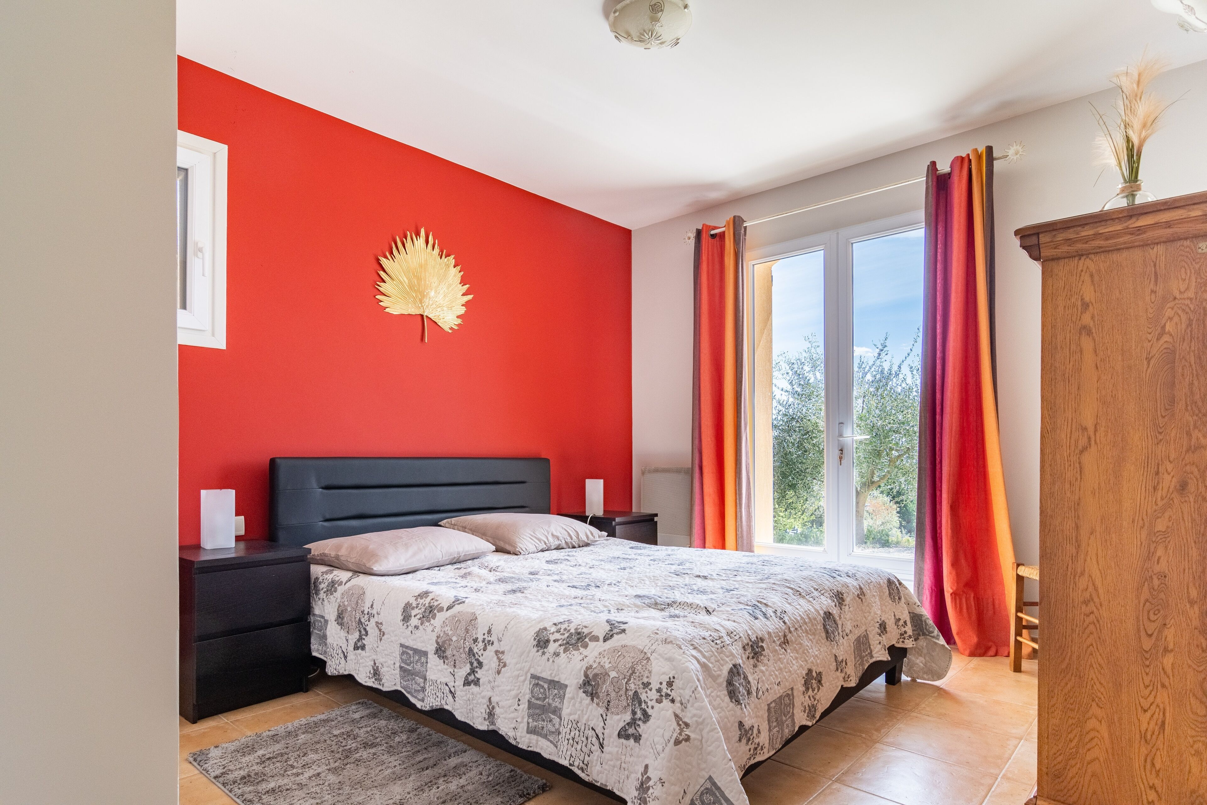 Een slaapkamer met een rode wand, een bed met beddengoed met een patroon, een nachtkastje, een houten garderobe en een raam met gordijnen.