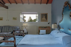 Un dormitorio con cama, una silla, un escritorio, un ventanal y un cuadro en la pared.