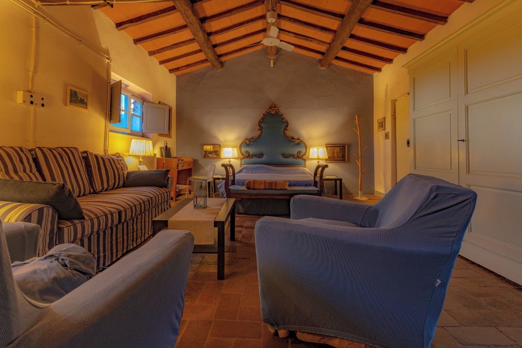A cozy room with a bed, a sofa, and two armchairs. There is a wooden ceiling and a white door in the background.