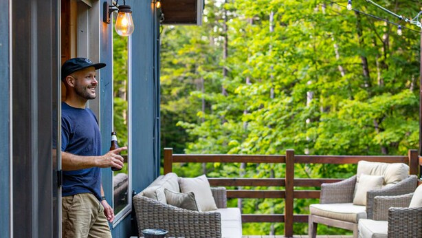 A man standing at a wooden deck door with wicker furniture and string lights.