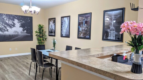 A modern kitchen with a granite countertop, black chairs, and a dining table. There are framed cityscape pictures on the wall and a potted plant on the counter.