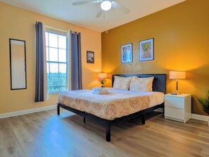 A bedroom with a bed, two bedside tables, a ceiling fan, a window with curtains, and wall-mounted artwork. - Kingsize 3BR Fast Wifi Free Parking Near Disney/Universal. (Kissimmee)