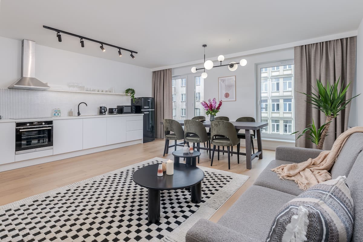 Un appartement moderne comprenant une cuisine, un coin repas et un salon. Il est doté d’un tapis à motifs noir et blanc, d’un canapé gris et d’une table à manger avec des chaises.