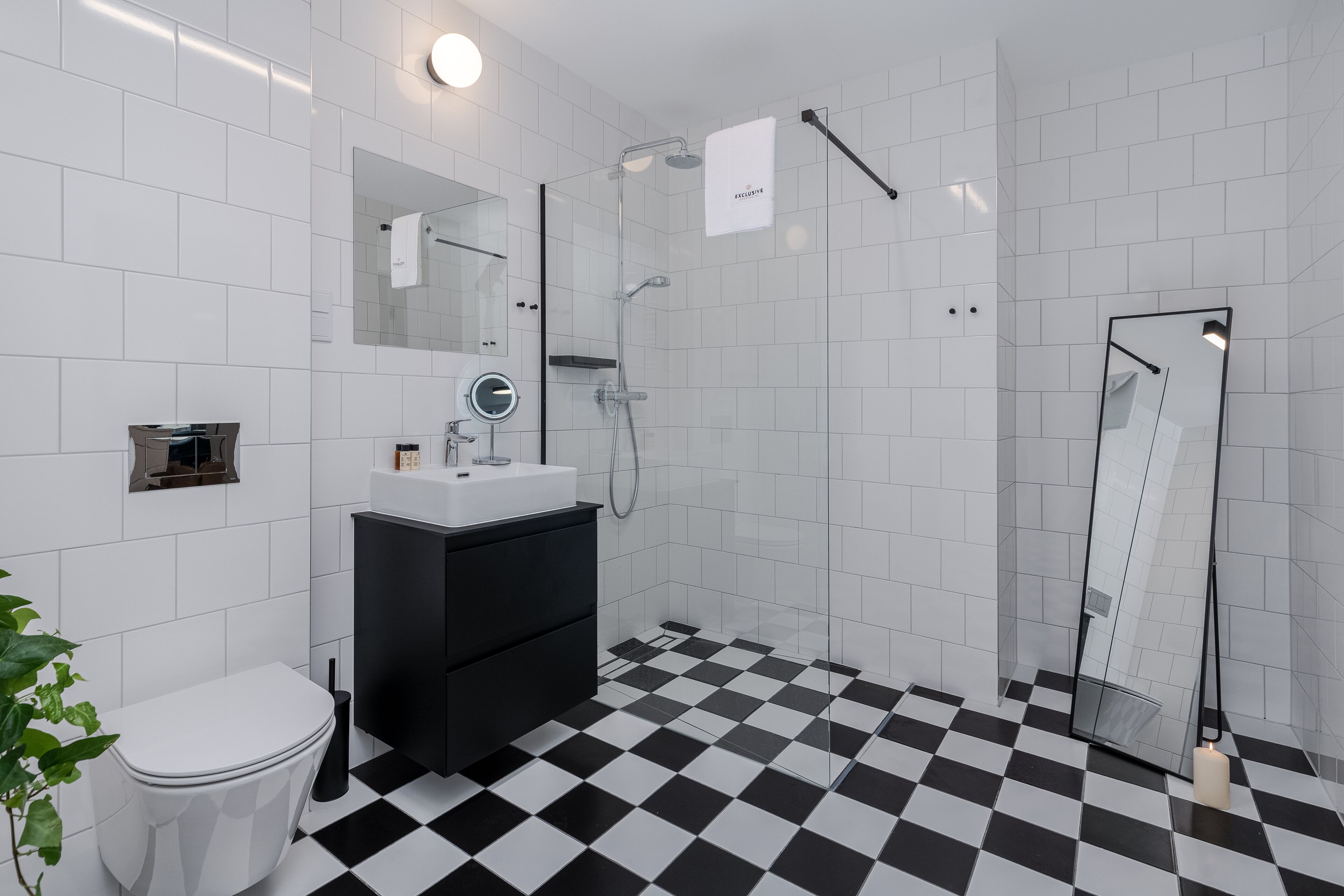 Een badkamer met een zwart-wit schakeerd motief op de vloer, een wit toilet, een zwart aanrecht met een spiegel en een douchecabine met glazen wanden.