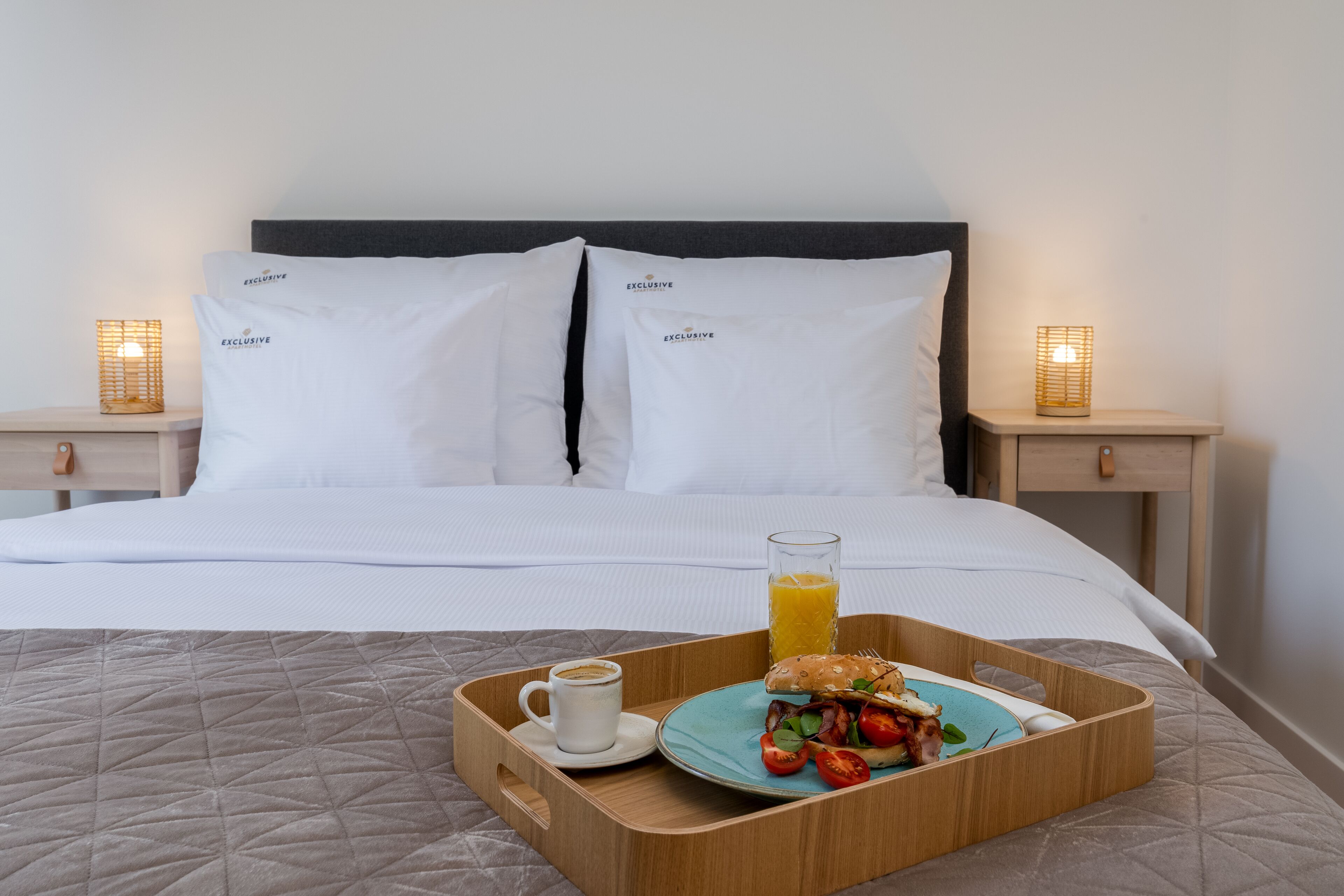 A neatly made bed with white bedding and two pillows labeled "EXCLUSIVE". A wooden tray with a breakfast plate, a glass of orange juice, and a cup of coffee is placed on the bed.