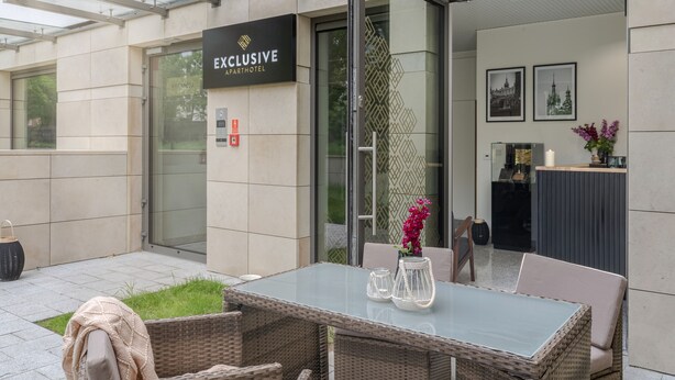 Ein Sitzbereich im Freien mit Glastisch und Korbstühlen, ein Schild mit der Aufschrift 'Exclusive Apartment Hotel' und einer Wand mit Muster.