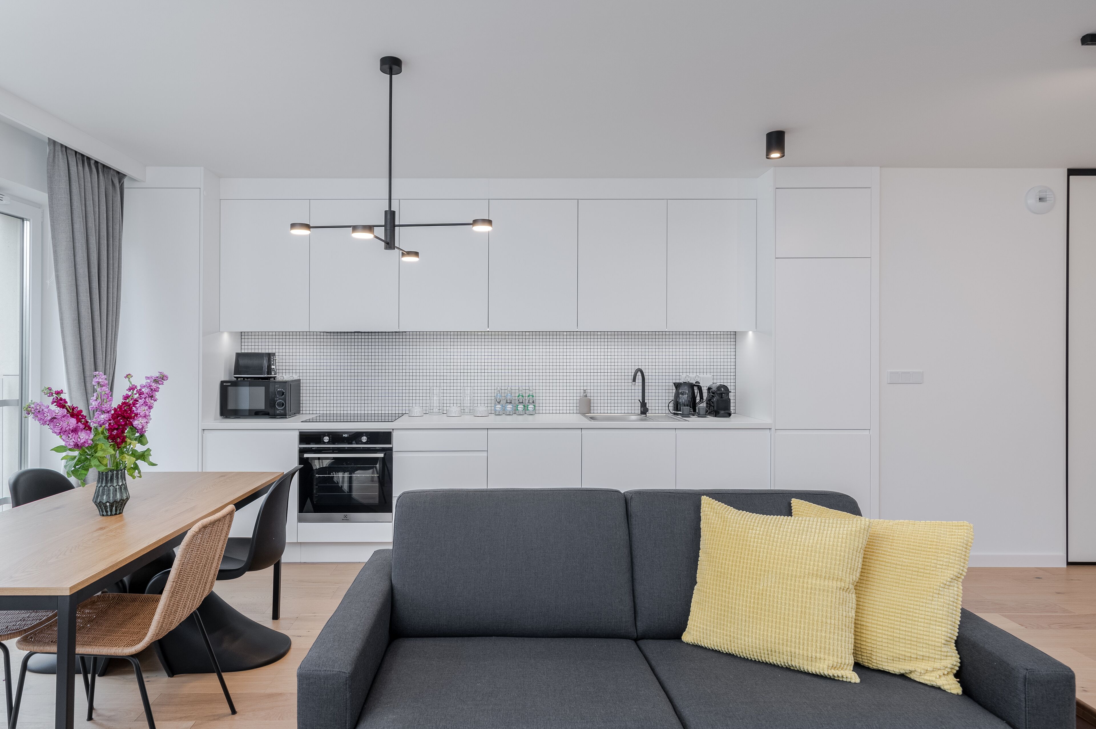 Eine moderne Küche mit weißen Schränken, ein Essbereich mit einem Holztisch und -stühlen sowie ein graues Sofa mit gelben Kissen.