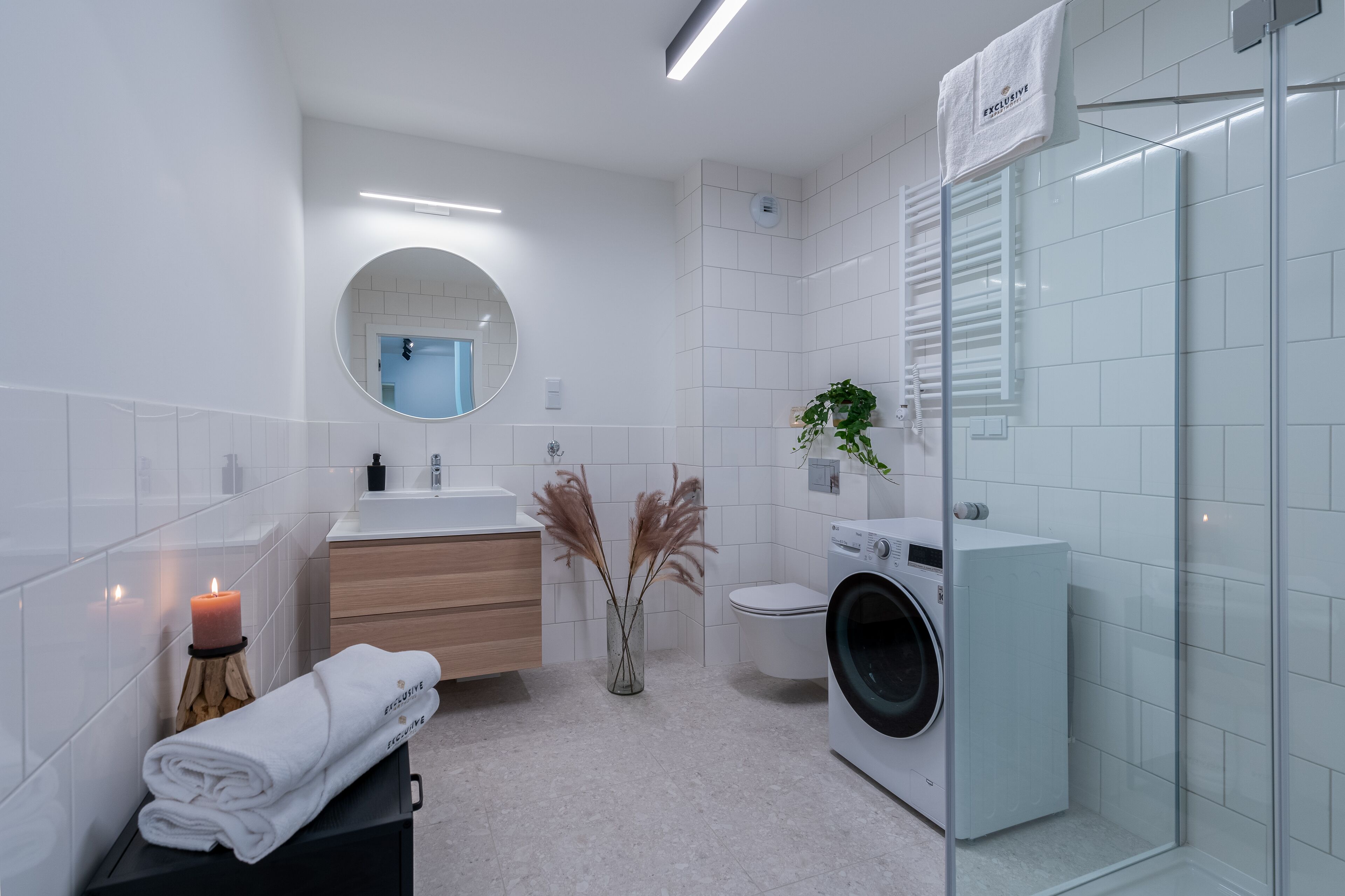 Een moderne badkamer met een ronde spiegel, witte tegels, een wastafel, een toilet, een wasmachine en een kaars.