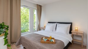 Ein ordentlich gemachtes Bett mit einem Frühstücksbrettchen, ein Nachttisch mit Lampe und ein Fenster mit Vorhängen.