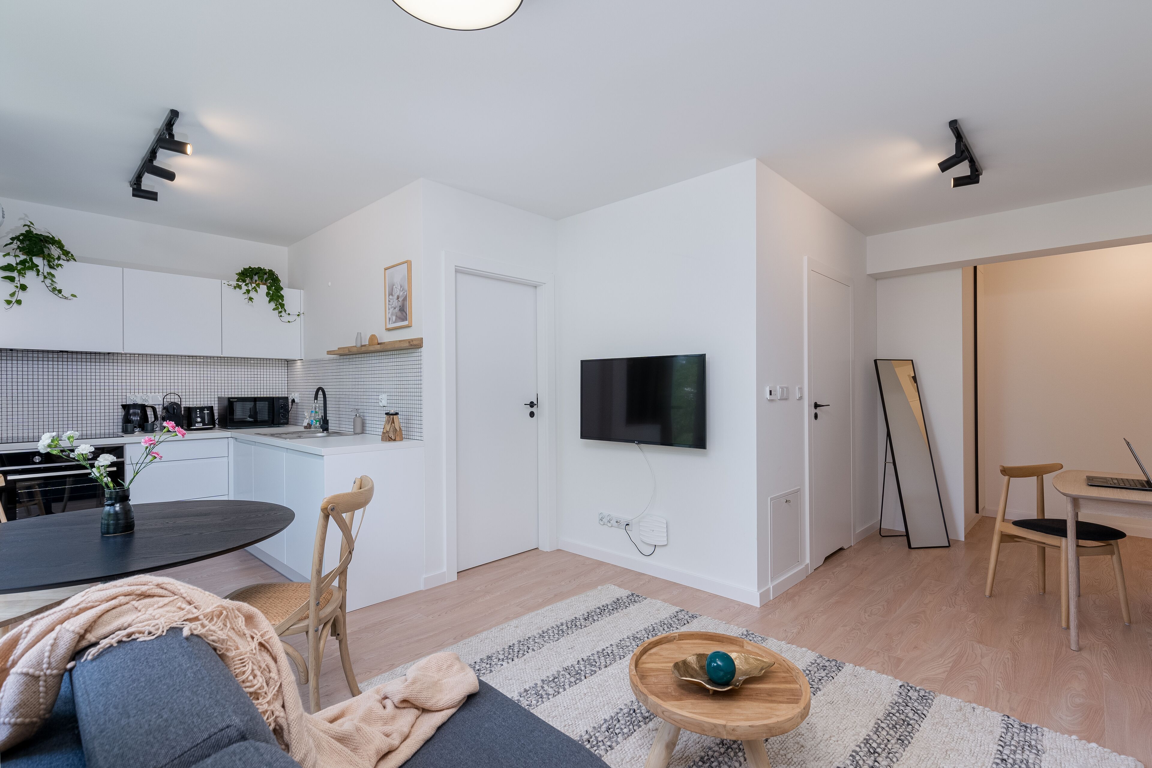 Una cuina moderna amb armaris blancs, una zona de menjador amb una taula rodona i cadires, un televisor penjat a la paret i una petita taula auxiliar de fusta amb un bol.