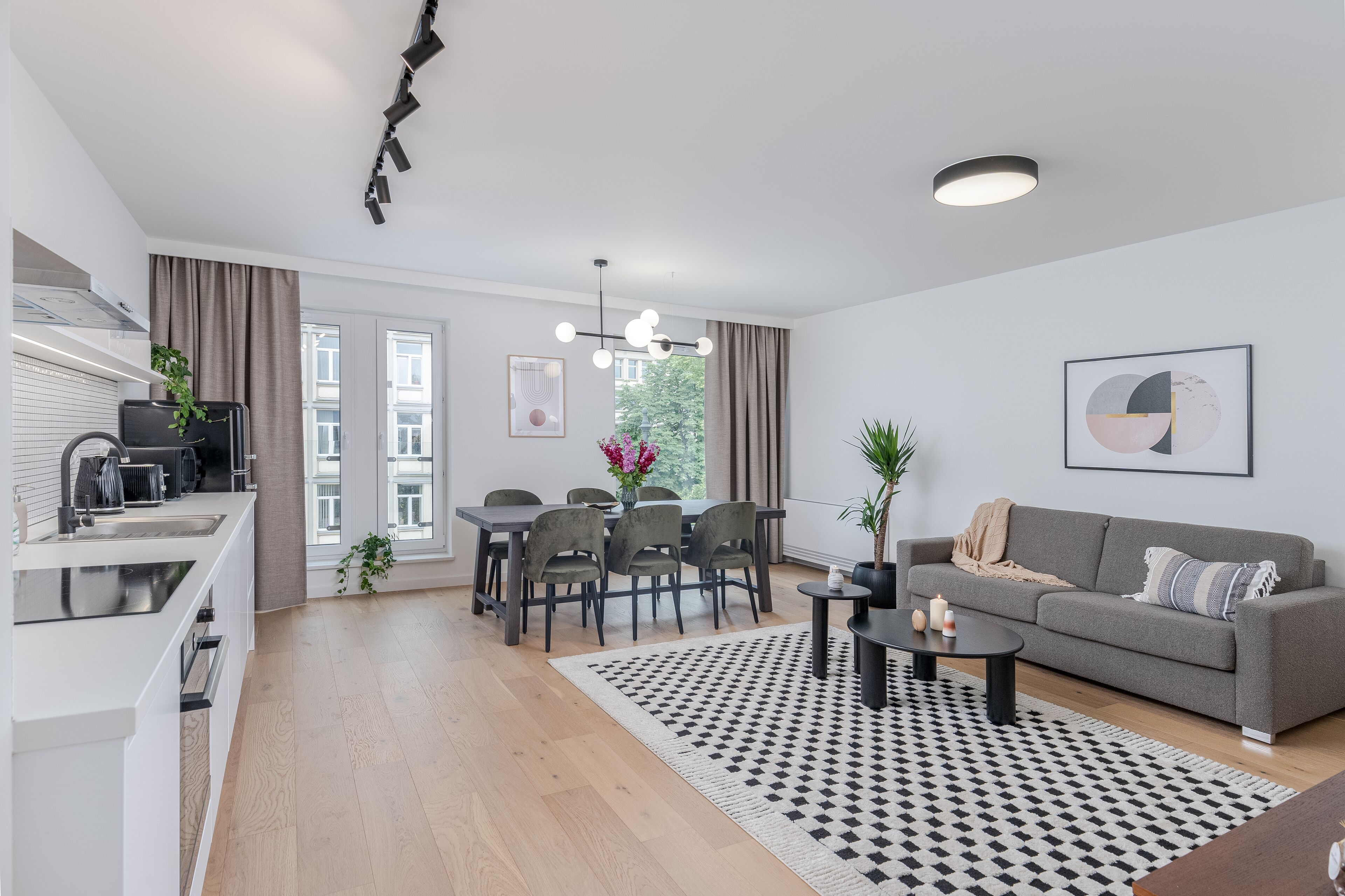 A modern apartment with a kitchen, dining area, and living room. It features a dining table with chairs, a sofa, and a coffee table. There is a window with curtains, a ceiling light, and a framed picture on the wall.
