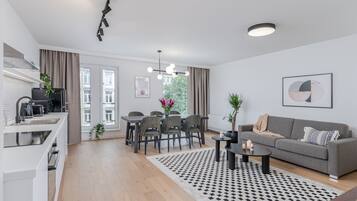 A modern apartment with a kitchen, dining area, and living room. It features a dining table with chairs, a sofa, and a coffee table. There is a window with curtains, a ceiling light, and a framed picture on the wall.