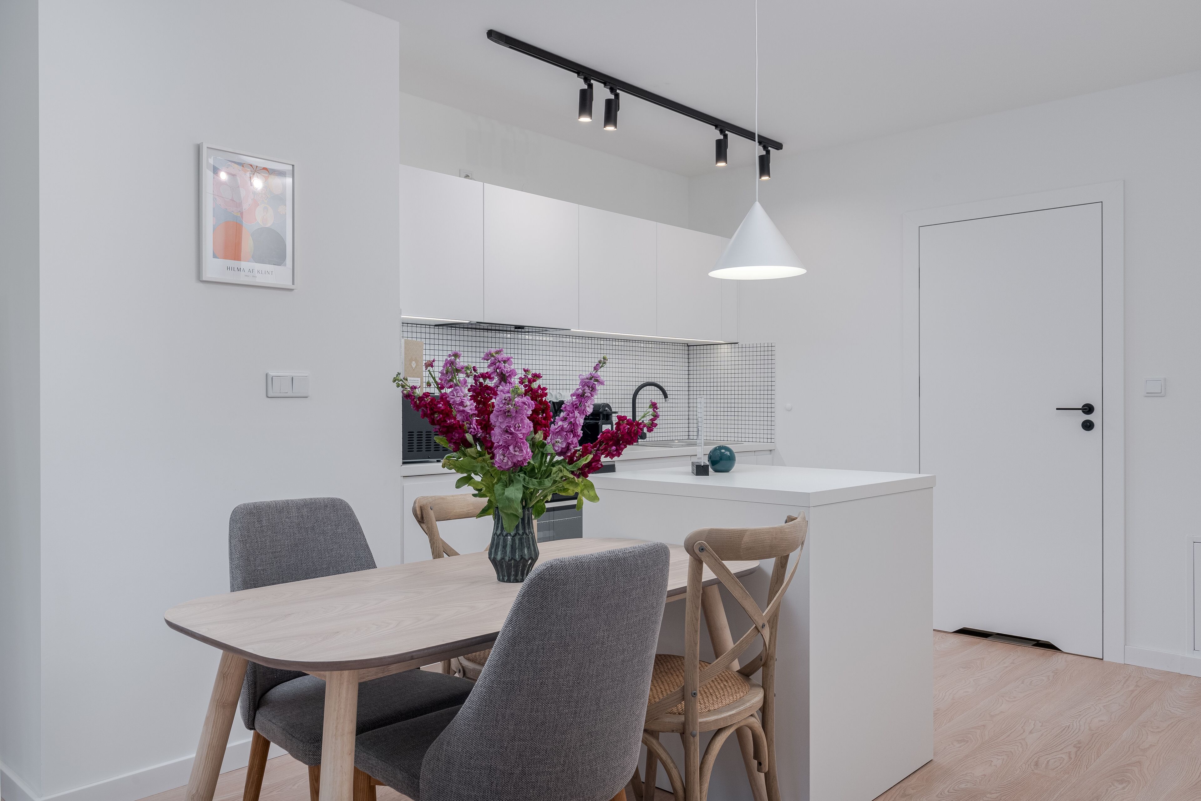 A modern kitchen with white cabinets, a wooden dining table, and a vase of purple flowers.