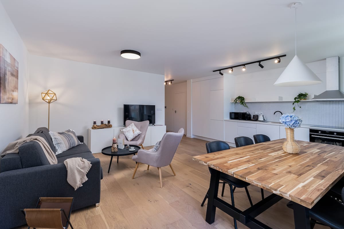 Un salon moderne comprenant un canapé, une table basse et des fauteuils, une table à manger en bois avec des chaises, un téléviseur et un coin cuisine avec des placards et un luminaire suspendu.