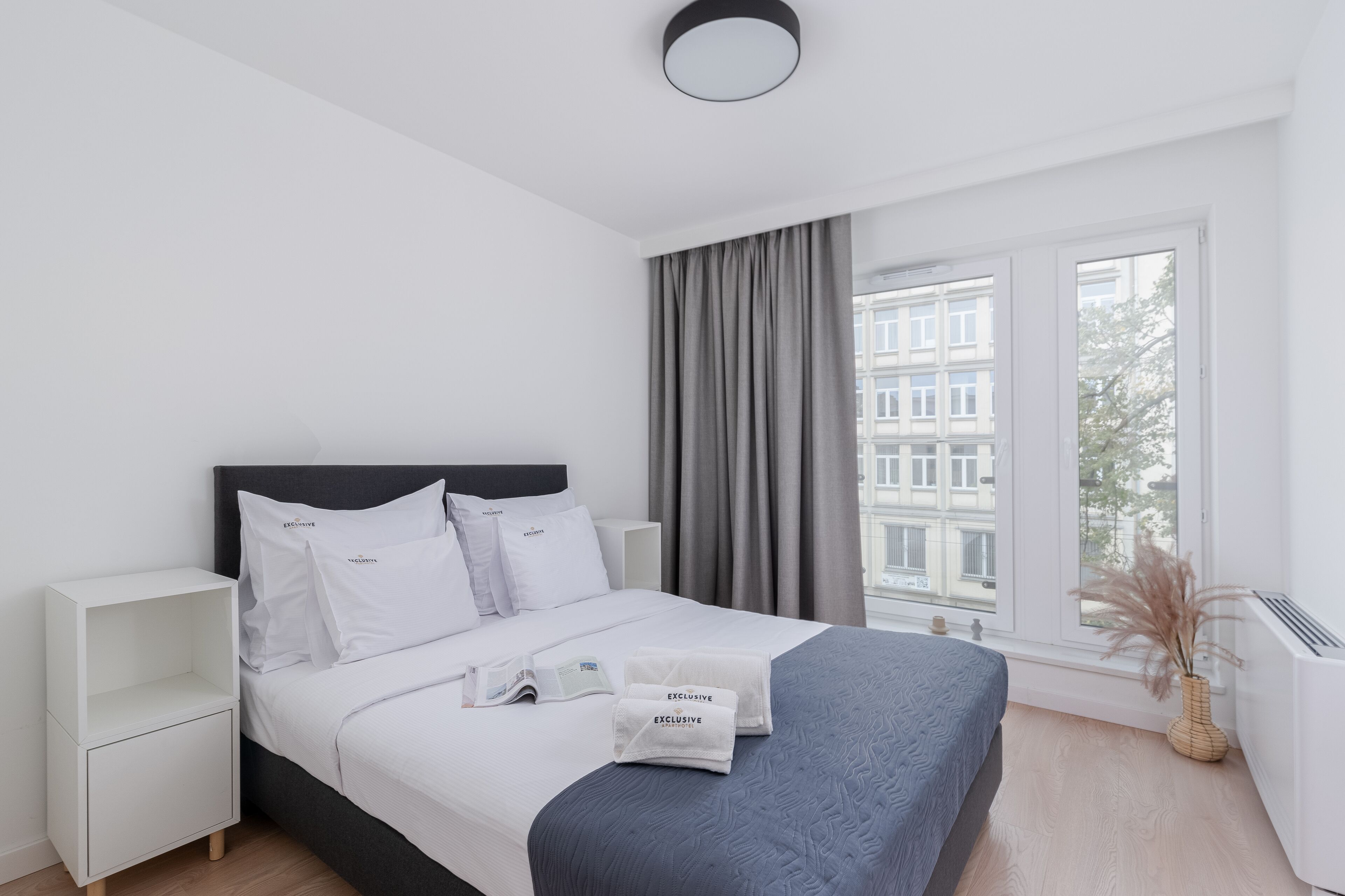 A modern hotel room with a bed, white nightstands, a window with curtains, and a view of buildings outside.